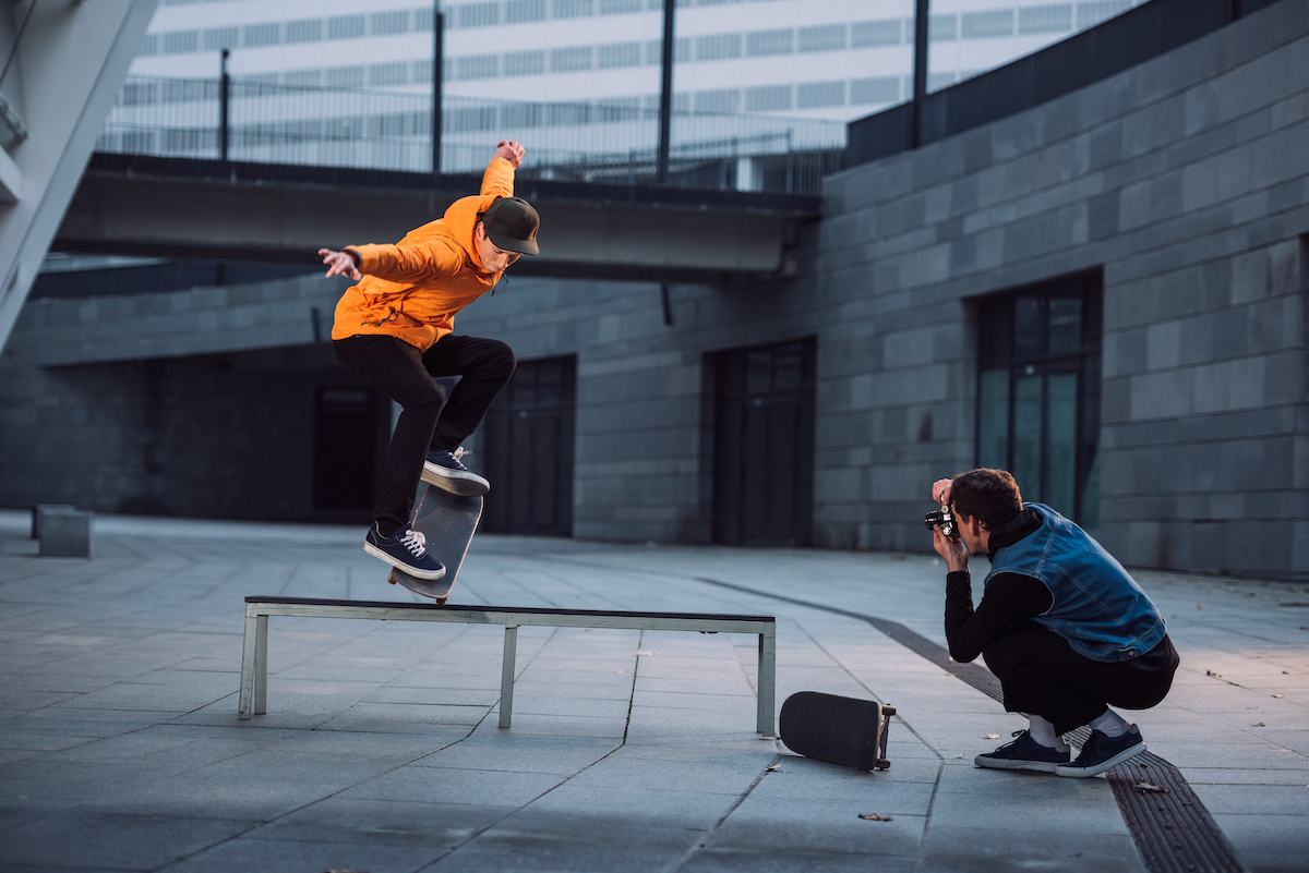 A man in a bright orange hoodie doing a skateboard trick while a photographer captures the shot.
