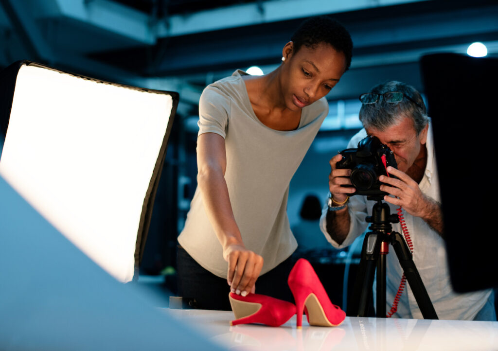 A woman placing red heels on a table during a retail store photography shoot and a man photographing them.