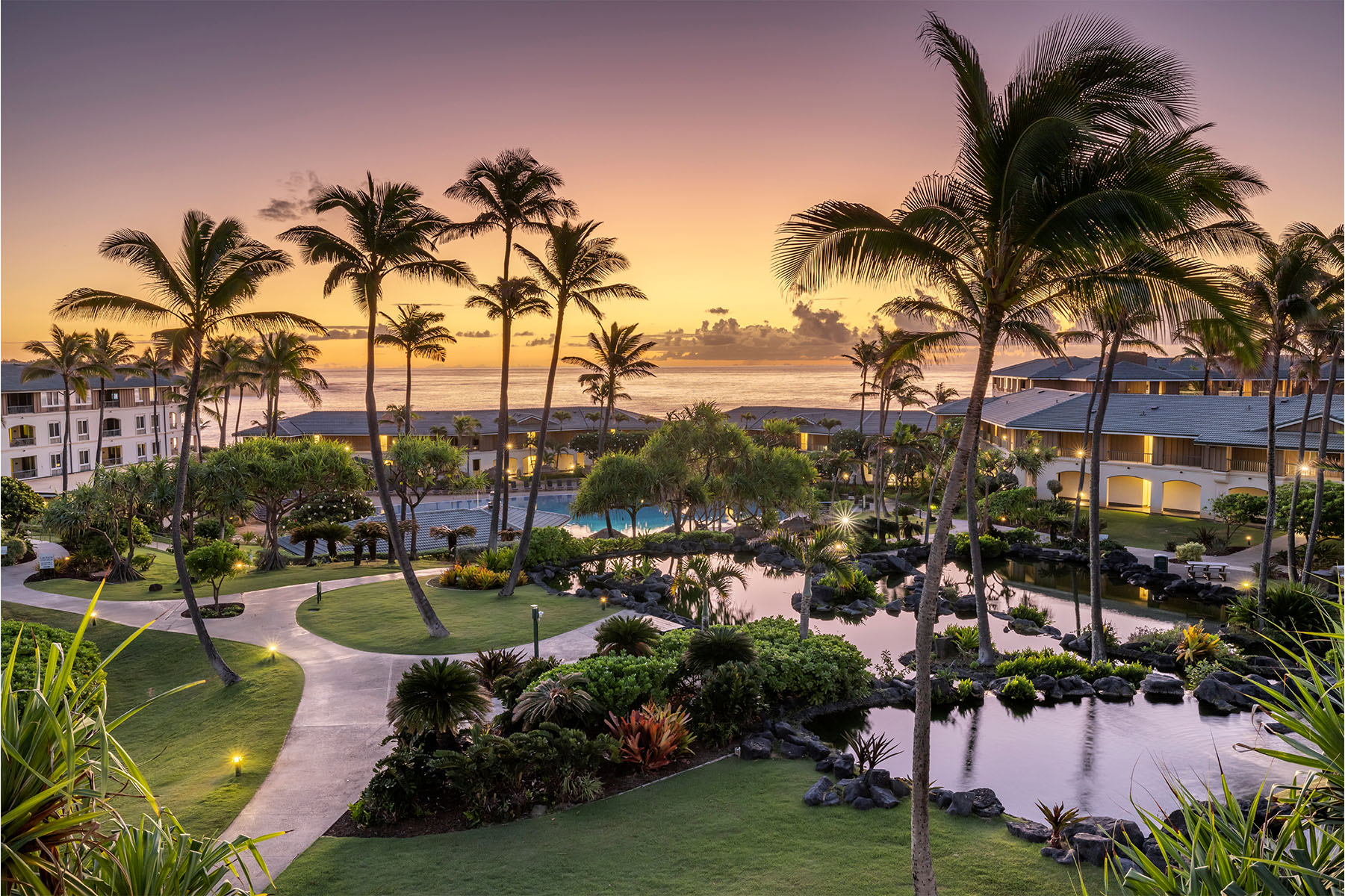 Scenic resort landscape at sunset with palm trees, walking paths, lush gardens and an ocean view in the distance.