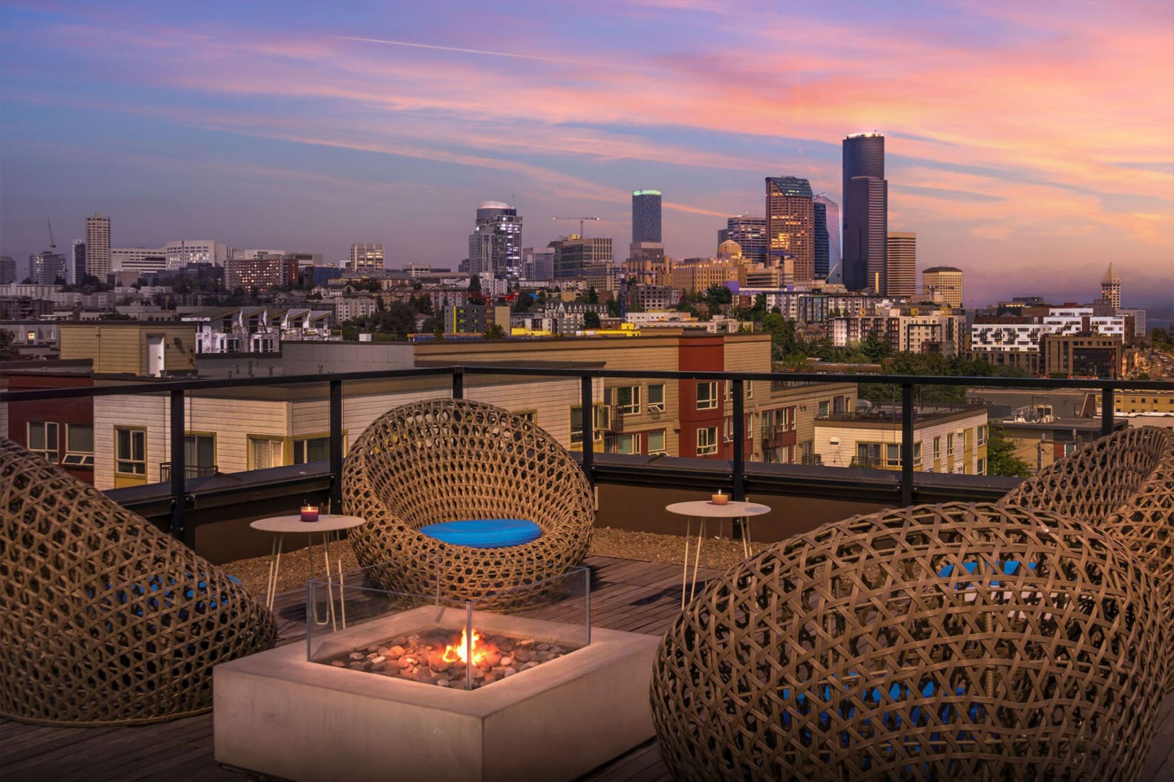 Rooftop lounge with woven seating, fire pit and skyline views of downtown buildings at dusk.