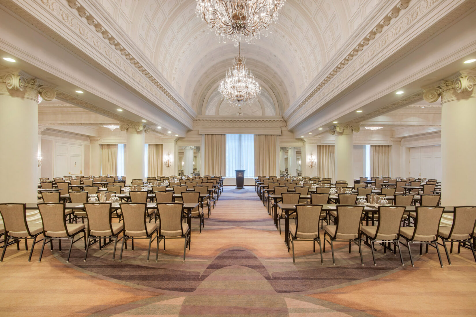 Grand hotel ballroom with ornate vaulted ceilings, tiered chandeliers and rows of chairs arranged classroom-style facing a podium.