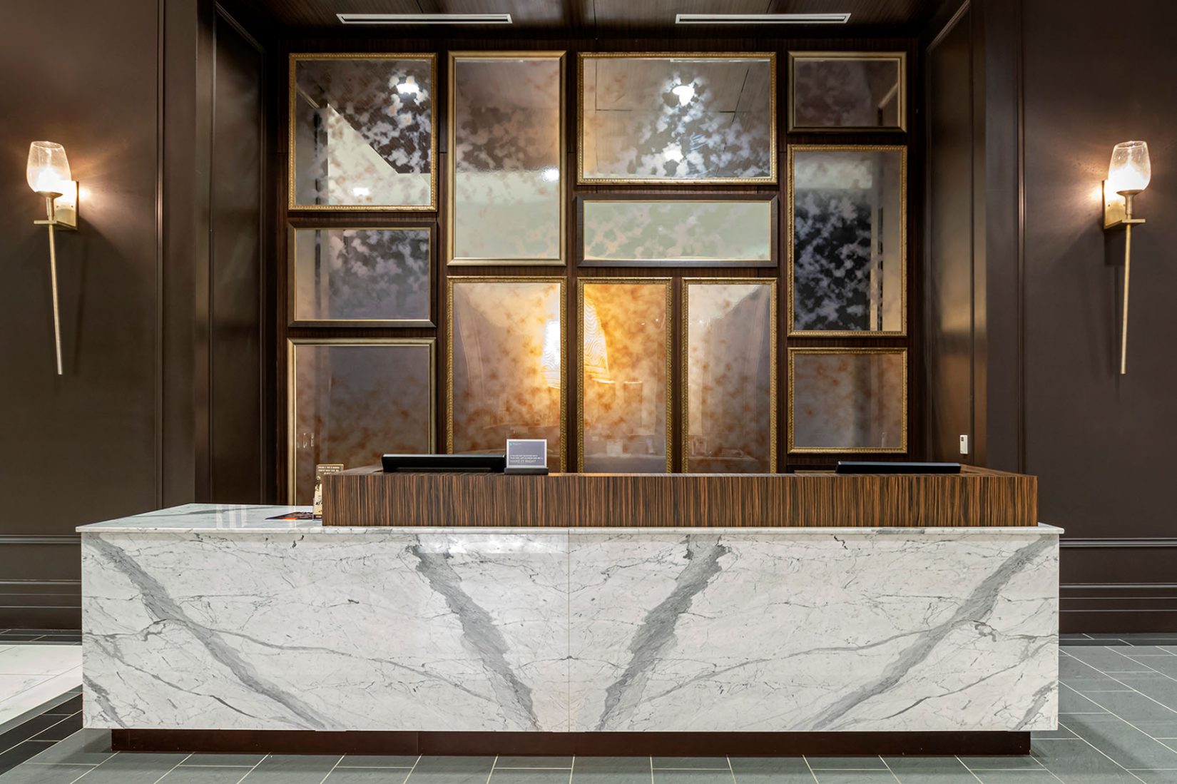 Elegant hotel reception area with a marble front desk, dark wood accents, wall-mounted decorative panels and warm ambient lighting.