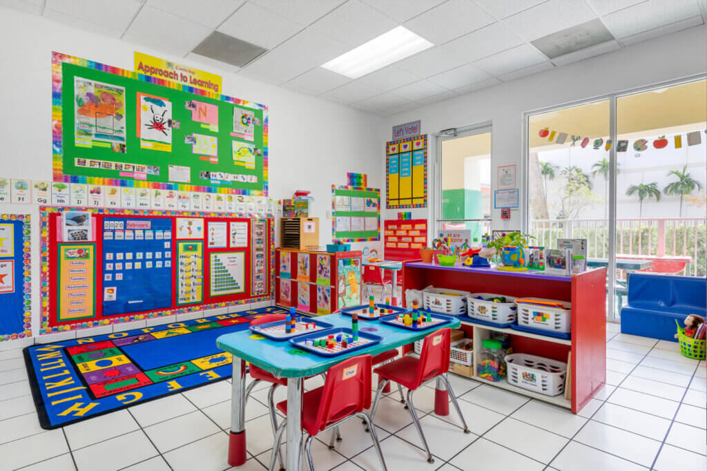 Bright early education classroom with colorful learning displays, a small activity table, children’s chairs, storage bins, and large windows letting in natural light.