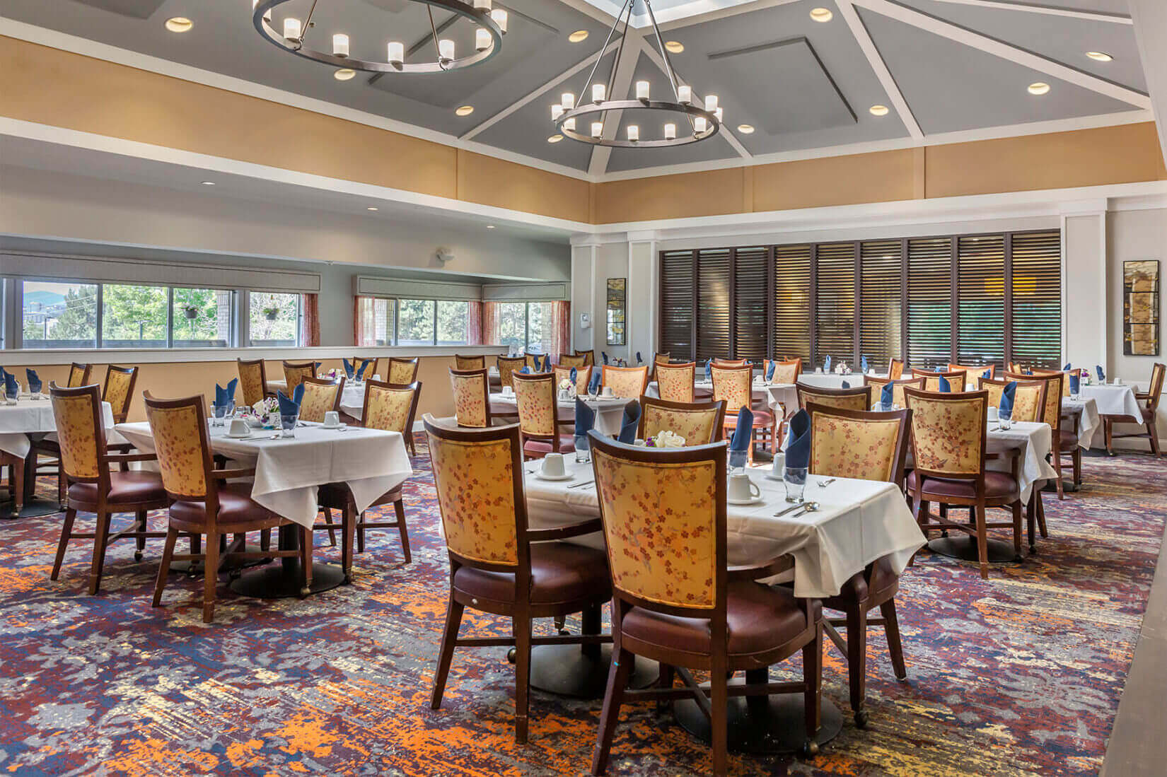 Senior living dining room with white tablecloths, set tables, patterned chairs and large windows.